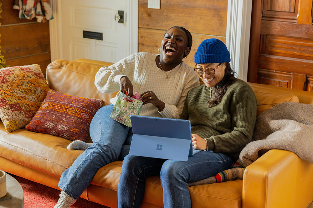 Two ladies sitting on a couch, looking at an ipad and laughing.