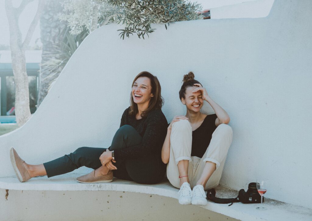 Two women laughing next to a wall with a camera and glass of wine at their side.