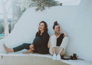 Two women laughing next to a wall with a camera and glass of wine at their side.