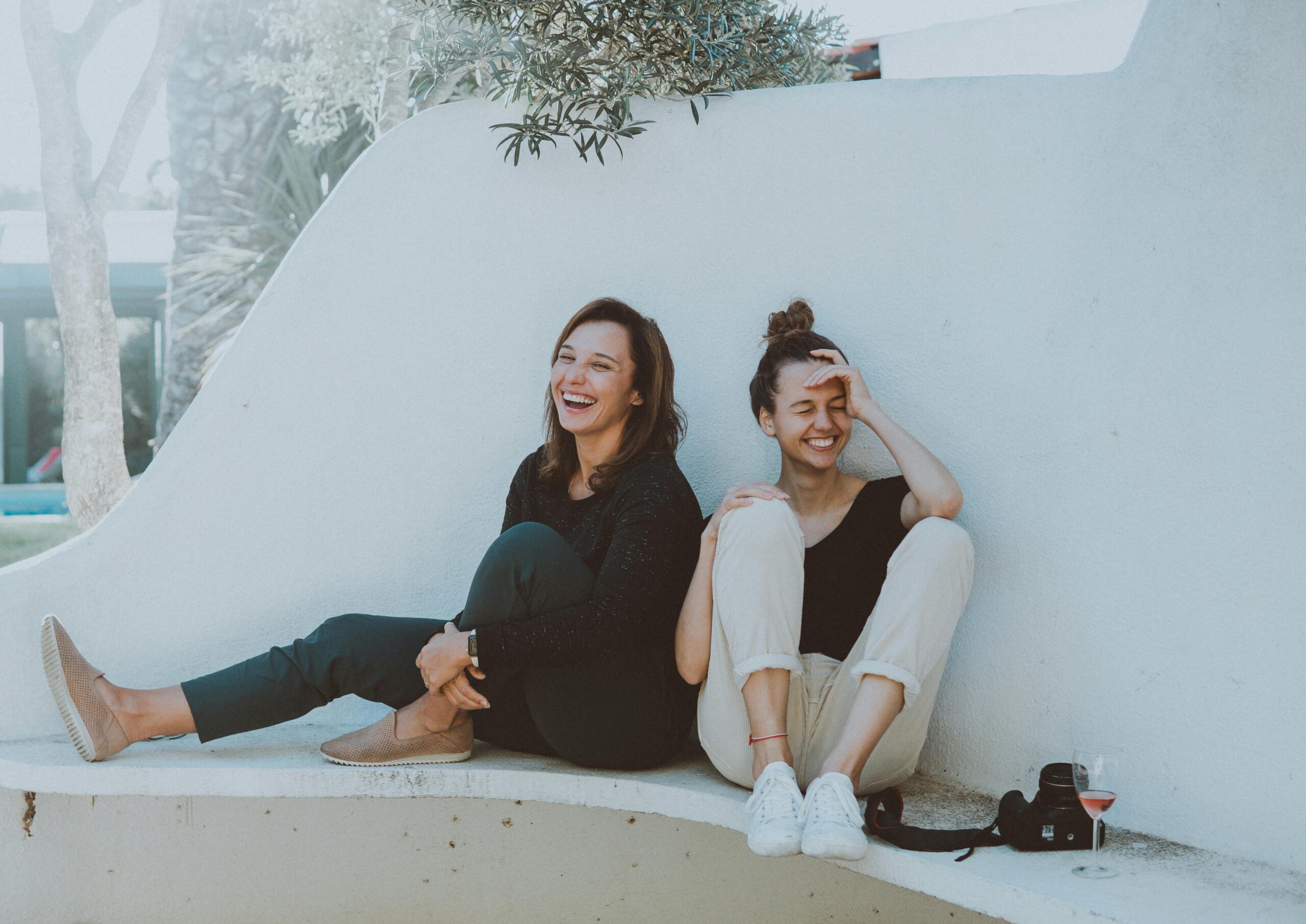 Two women laughing next to a wall with a camera and glass of wine at their side.