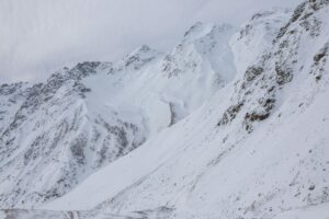 Expansive view of a rugged mountain range blanketed in snow, with sharp ridgelines and scattered rocky outcrops under an overcast sky.