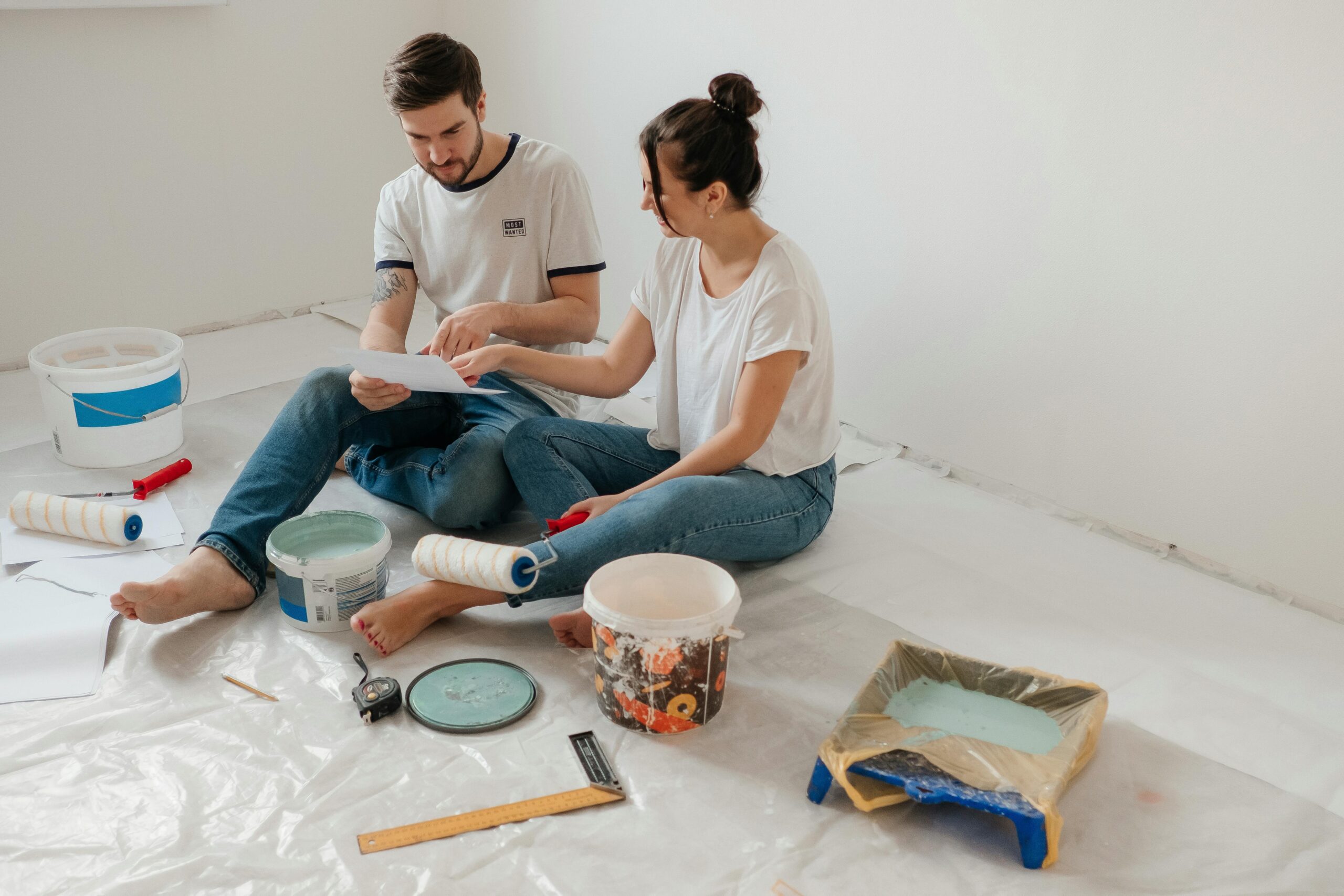 Young couple sitting on the floor of their home surrounded by paint supplies, reviewing renovation plans together while preparing to repaint a room.